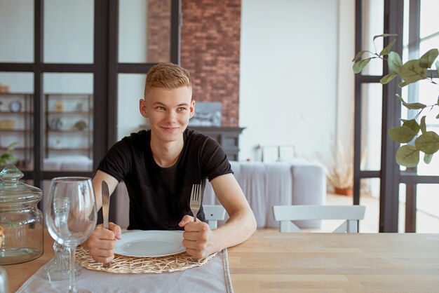 Hambriento joven guapo adolescente en camiseta negra sentado en la mesa con cuchillo tenedor