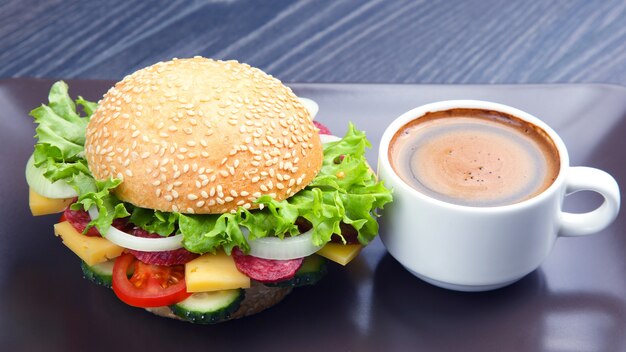 Hamburger mit Gemüse und Wurst und Kaffee auf grauem Hintergrund. Fast Food und Frühstück. Kalorien und Ernährung.