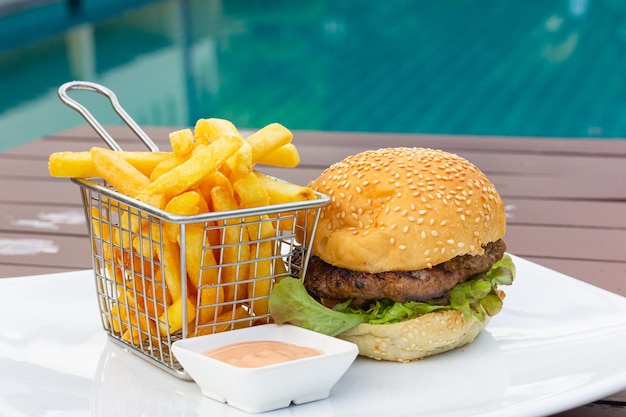 Hambúrguer clássico com cesta de aço inoxidável com batatas fritas e prato de molho rosa à beira da piscina