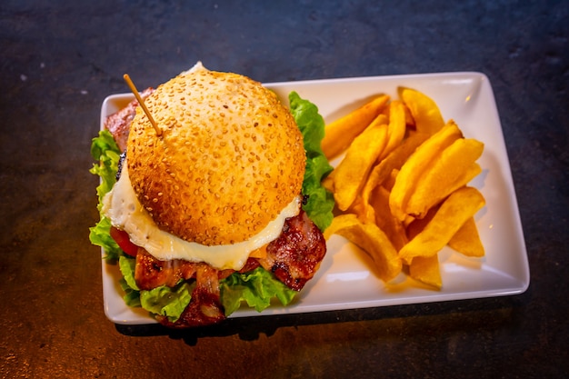 Hambúrguer com bacon e batata frita em um fundo preto, em um prato branco