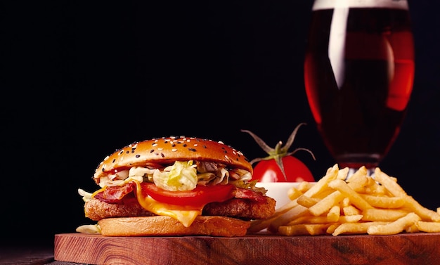 Hambúrguer com costeleta de frango, bacon frito e vegetais, batata frita, copo de cerveja em uma placa de madeira