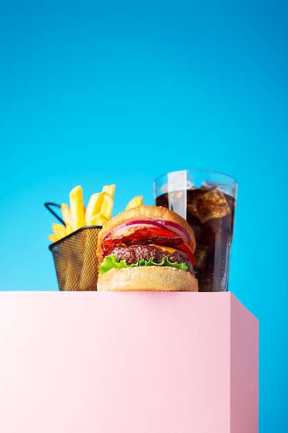 Hambúrguer de carne fresca e suculenta, coca-cola e batatas fritas fritas colocadas no carrinho rosa e no fundo azul. Copie o espaço para texto, visão de herói moderno