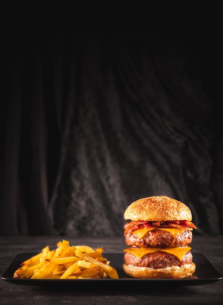 Hambúrguer duplo de carne e bacon com queijo cheddar e batatas fritas em mesa preta