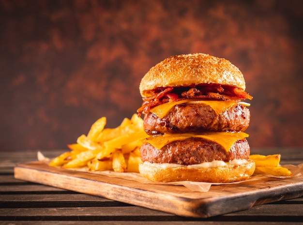 Hambúrguer duplo de carne e bacon com queijo cheddar e batatas fritas em uma tábua de madeira