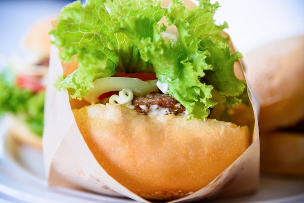 Hambúrguer é um fast food americano delicioso, closeup um hambúrguer feito de carne de porco ou vaca, pão de alface verde, cebola e tomate em um saco de papel e prato no fundo da mesa em casa para fazer um piquenique ou festa