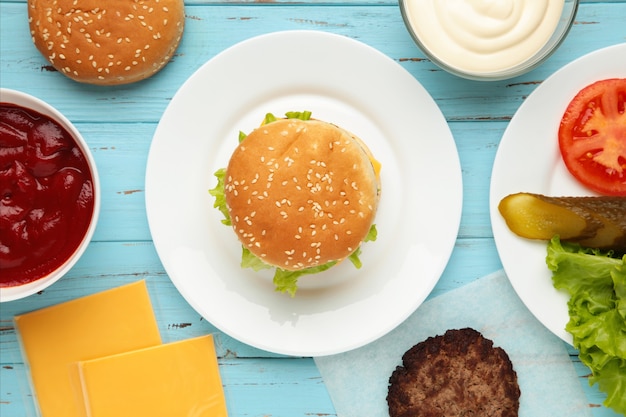 Hambúrguer em um prato branco e ingredientes na mesa azul.
