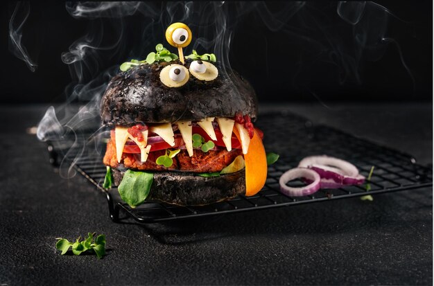 Foto hambúrguer monstro com pão preto, queijo patty vegano, olhos de azeitona e fumaça no fundo preto comida de halloween