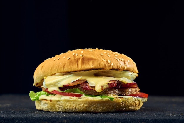 Hambúrguer suculento com ovo, legumes, bacon e molho em um fundo escuro.
