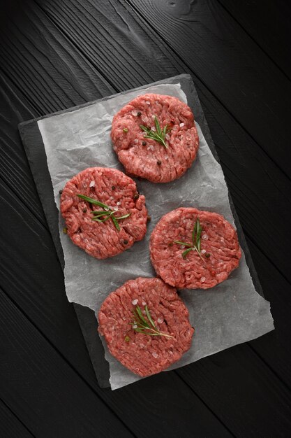 Hambúrgueres de carne crua na tábua de madeira do açougueiro, alecrim e pimenta fundo preto vista de cima