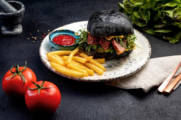 Hamburguesa con bollos negros con bistec y verduras servida con papas fritas y salsa sobre un fondo oscuro