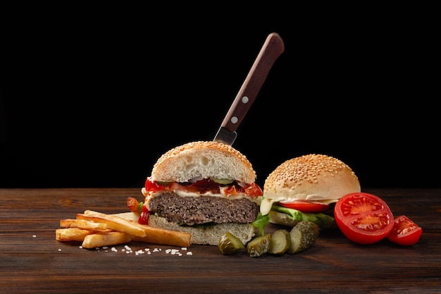 Hamburguesa casera cortada en medio primer plano con carne de res, tomate, lechuga, queso y papas fritas en la mesa de madera. En la hamburguesa metió un cuchillo