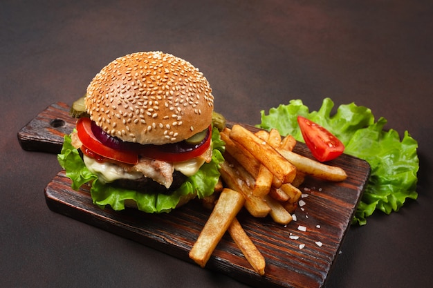 Hamburguesa casera con ingredientes de carne de res, tomates, lechuga, queso, cebolla, pepinos y papas fritas en la tabla de cortar y fondo oxidado