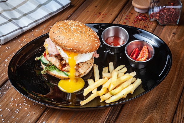 Hamburguesa fresca con huevo de gallina, lechuga y salsa en un plato negro con papas fritas. Comida rápida estadounidense. Chickenburger con copia espacio sobre fondo de madera. De cerca, enfoque selectivo. comida. Menú de parrilla