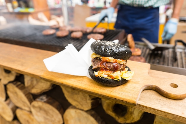 Foto hamburguesa gourmet servida sola en una tabla de cortar de madera con servilletas en el mostrador del puesto de comida con una persona cocinando empanadas en la parrilla en segundo plano.
