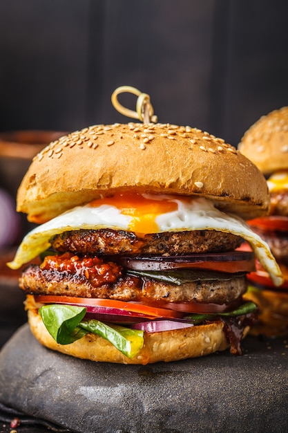 Foto hamburguesa hecha en casa de la carne con el huevo, la salsa y las verduras en fondo oscuro.