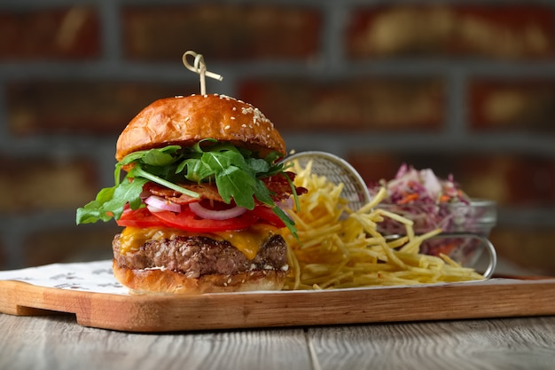 Hamburguesa en la mesa de madera con queso, tocino, tomates, ensalada verde y roja y papas fritas