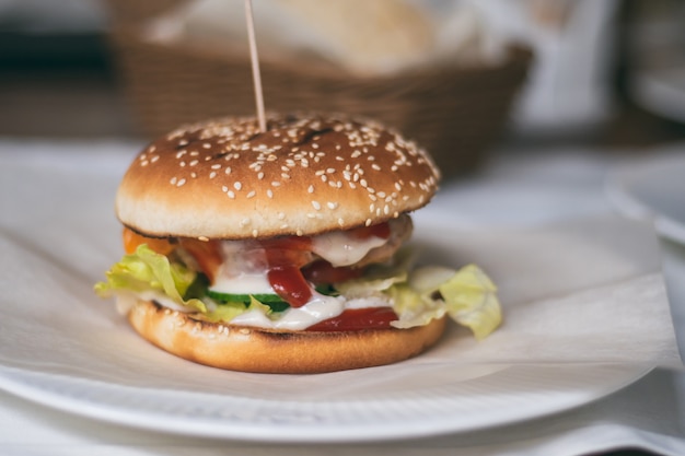 Hamburguesa parcialmente borrosa unida con un pincho de madera en un plato. Bollo de sésamo, chuleta, pepino y tomate, lechuga, salsa