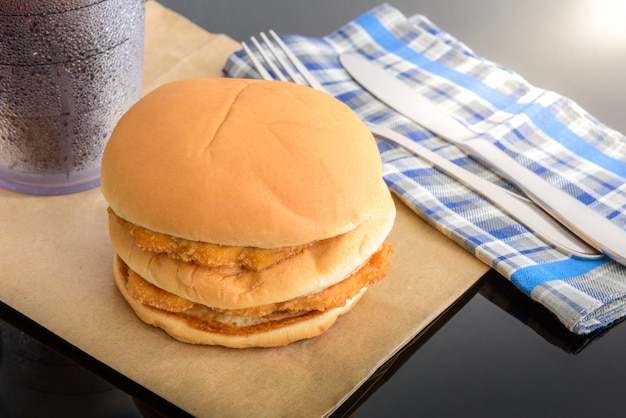 hamburguesa de pollo y vaso de cola en papel marrón con cuchillo y tenedor en la mesa superior, reflexiones