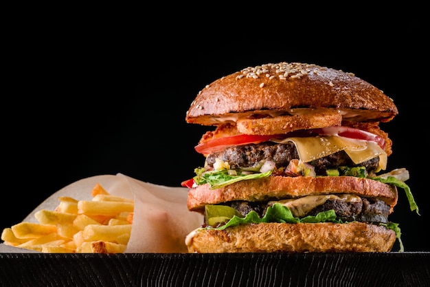 Hamburguesa de queso con carne a la parrilla, queso, tomate y patatas sobre superficie de madera oscura. Plantilla de comida rápida. Foto real. Ideal para publicidad. De cerca. Bodegón hamburguesa y papas fritas