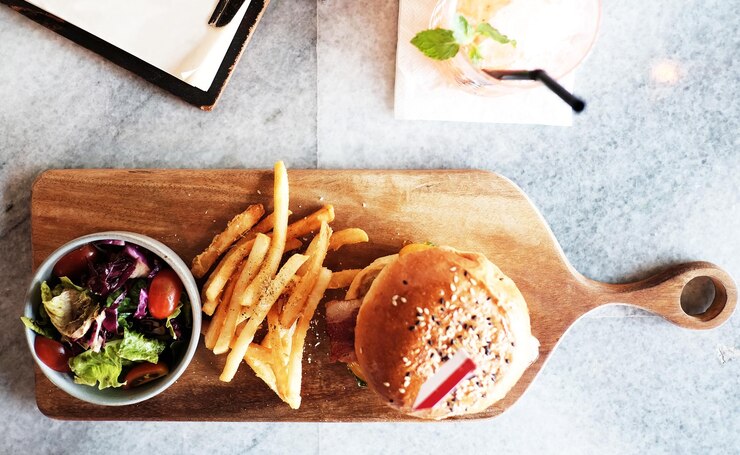 Hamburguesa de queso con jamón servida con un plato de ensalada y papas fritas colocadas en una tabla de cortar de madera