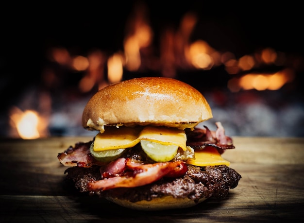 Una hamburguesa con queso, tocino y pepinillos descansa sobre una tabla de madera frente a una chimenea.