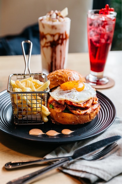 La hamburguesa sabrosa con el huevo frito sirvió con las fritadas en placa negra en la tabla de madera.