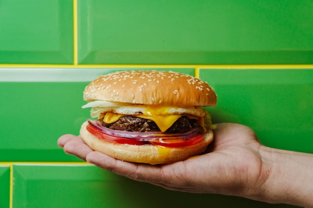 Hamburguesa de ternera con queso y verduras en mano de hombre en pared verde