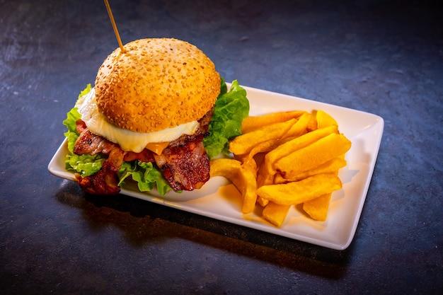 Hamburguesa con tocino y papas fritas sobre un fondo negro, sobre una placa blanca.