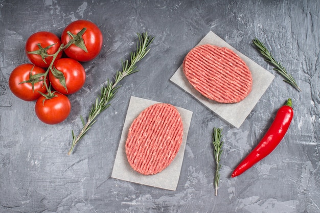 Hamburguesas crudas en el papel de pergamino con los tomates, las pimientas de chile y el romero.