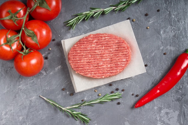 Hamburguesas crudas en el papel de pergamino con los tomates, las pimientas de chile y el romero.