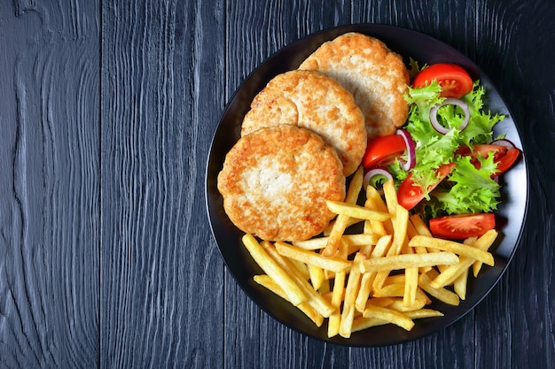 Foto hamburguesas de pavo con ensalada de tomate y lechuga y patatas fritas gruesas en una placa negra sobre una mesa de madera, vista desde arriba, flatlay, espacio libre para texto