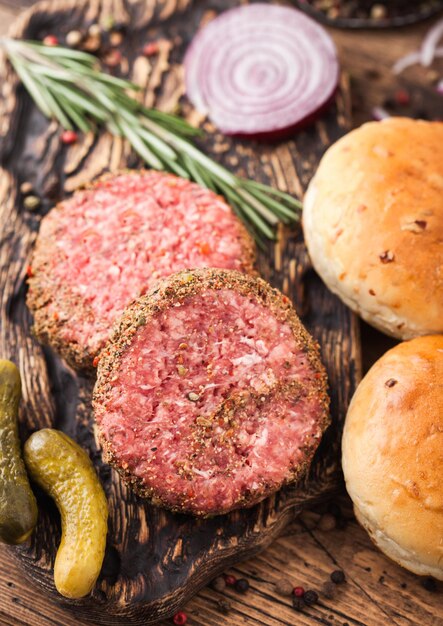 Hamburguesas de ternera con pimienta picada cruda fresca en una tabla de cortar vintage con bollos de cebolla y tomates sobre fondo de madera