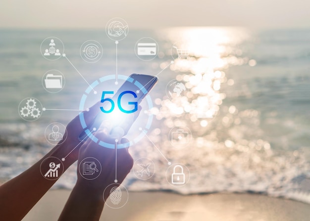 Hand der Frau, die das Mobiltelefon verwendet, verbindet das Netzwerk 5G mit dem Symbol am Strand Technologienetzwerk drahtlose Systeme und Internet der Dinge Weltweites Geschäftskonzept für mobiles Highspeed-Internet