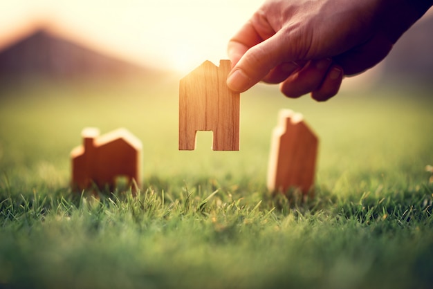 Hand der Frau, die Mini-Holzhausmodell auf grünem Gras wählt, Planung kaufen Immobilien, Öko-Hausikonenkonzept.