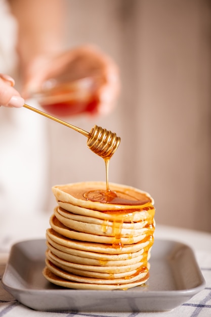 Hand der jungen Hausfrau, die frischen Honig vom Schöpflöffel auf Stapel des Stapelns der appetitlichen hausgemachten Pfannkuchen auf Teller kocht, der zum Frühstück gekocht wird