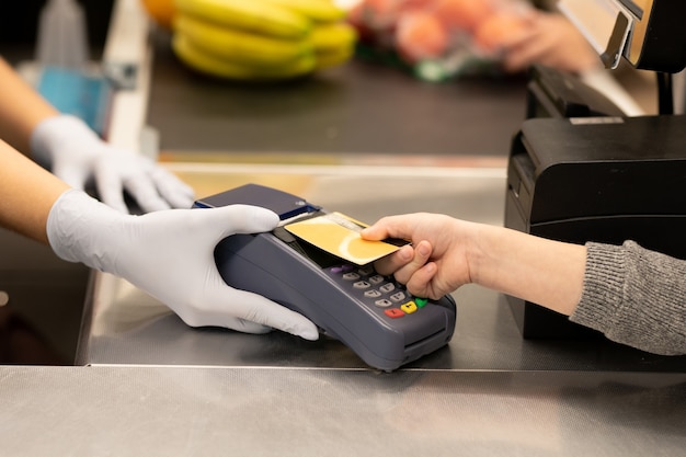 Hand der jungen Verbraucherin, die Kreditkarte über dem Bildschirm des Zahlungsterminals hält, der von einem behandschuhten Kassierer über der Theke im Supermarkt gehalten wird