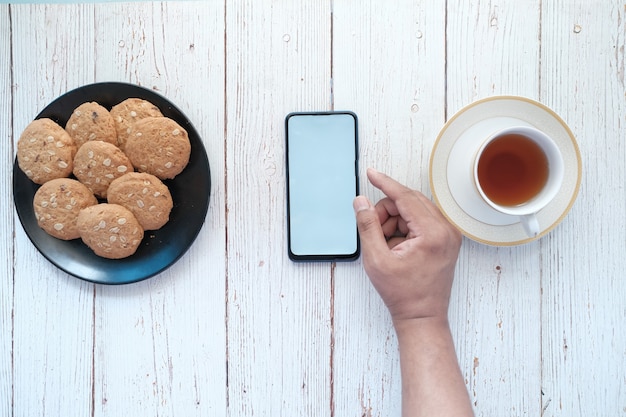 Hand des jungen Mannes mit Smartphone mit Tee und Keksen auf dem Tisch