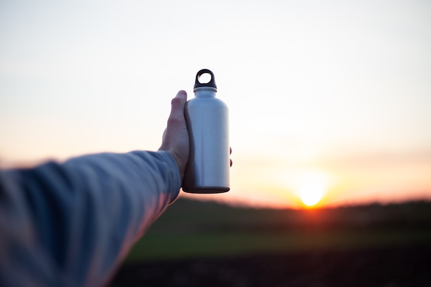 Hand des Mannes, der Aluminiumflasche für Wasser hält, auf Hintergrund des Sonnenuntergangs