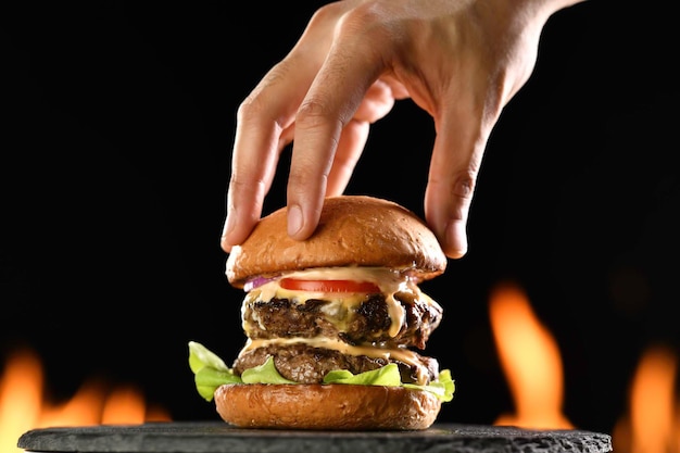 Hand, die das obere Burgerbrötchen auf einen doppelten Fleisch-Cheeseburger mit Gemüse mit Sauce legt, die auf flammendem Hintergrund sickert und heraustropft