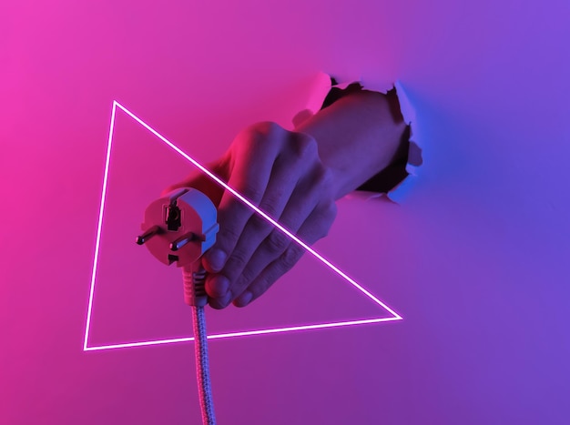 Hand, die den elektrischen Stecker durch ein zerrissenes Loch hält. Kreative Pop-Art-Rosa-Blau-Neonfarbe. Trendige Farbverlaufsbeleuchtung. Nachtlicht