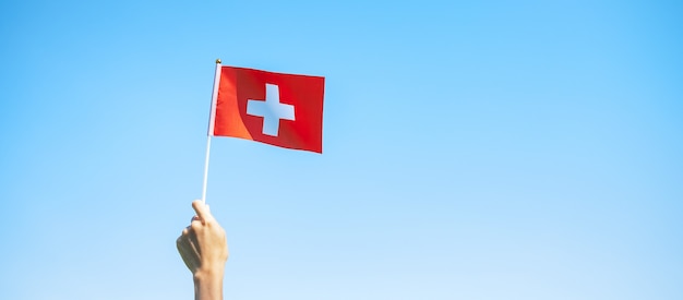 Hand, die die Schweiz-Flagge auf Hintergrund des blauen Himmels hält. Schweizer Nationalfeiertag und fröhliche Feierkonzepte