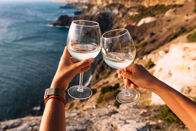 Hand, die ein Glas Wein über dem Meer hält Kinn-Kinn. Romantischer Urlaub. Zwei Hände, die Weingläser gegen das Meer halten. Platz kopieren