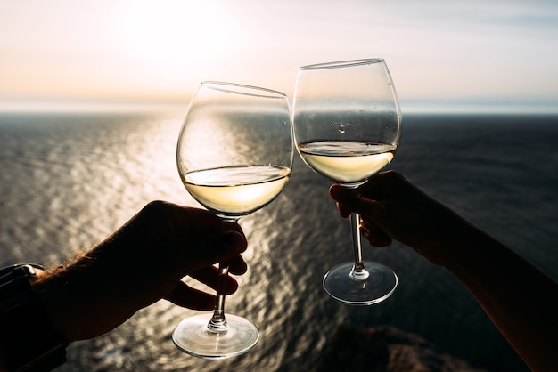 Hand, die ein Glas Wein über dem Meer hält Kinn-Kinn. Romantischer Urlaub. Zwei Hände, die Weingläser gegen das Meer halten. Platz kopieren