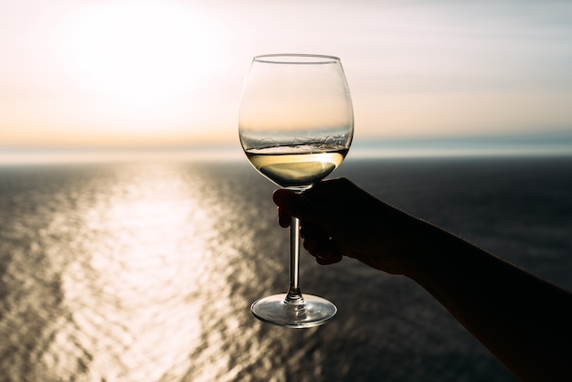 Hand, die ein Glas Wein über dem Meer hält Romantischer Urlaub. Hand, die ein Glas Wein gegen das Meer hält Den Sonnenuntergang mit Wein am Meer erleben. Platz kopieren