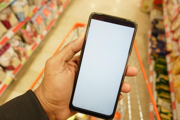 Foto hand, die ein handy mit weißem bildschirm hält, während sie im supermarkt einkaufen