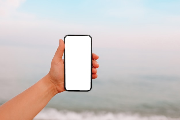 Hand, die ein leeres Smartphone am Strand mit dem Meer im Hintergrund zeigt. White-Screen-Mock-up