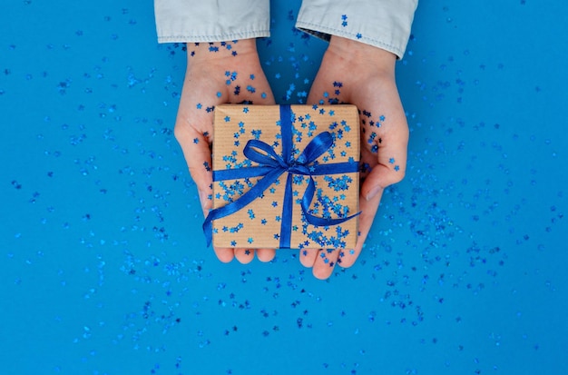 Foto hand, die eine geschenkbox mit blauem band auf blauem papierhintergrund gibt geburtstags- oder weihnachtsfeiertagskonzept