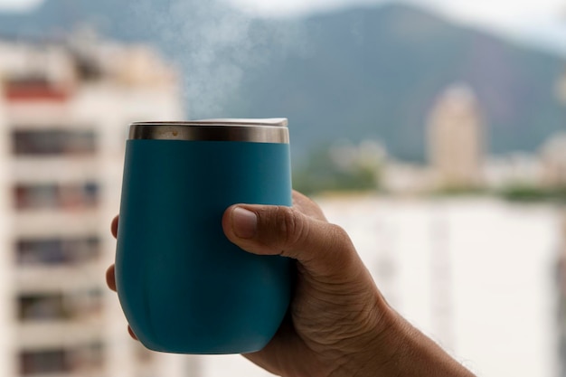 Hand, die eine Kaffeetasse hält, die aus Rauch mit Hintergrund der städtischen Umgebung kommt