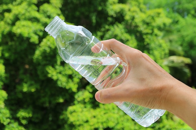 Hand, die eine Plastikflasche des Trinkwassers gegen grünes Laub hält