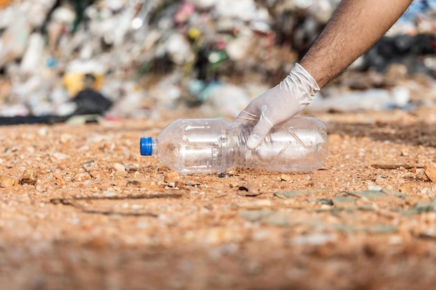 Hand, die eine Plastikflasche vor einem großen Berg und einem Müllhaufen hält. Abfallkunststoffe aus Industrie und Kommune. Weltumwelttag.